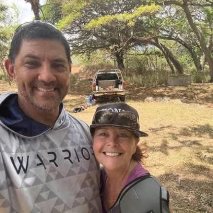 Ka'ala Souza and a companion smiling outdoors on the land in Moloka'i, with a pickup truck and large green trees in the background.